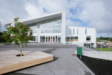A modern, architectural school building with a glass facade framed in white. An open courtyard is shown in the foreground
