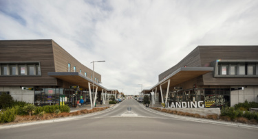 A street view framed by modern retail buildings clad in a wooden finish, people shop happily along the boardwalks.